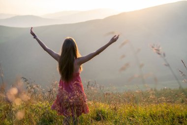 Kırmızı elbiseli genç mutlu kadın yolcunun arka görüntüsü. Çimenli bir yamaçta duruyor. Yazın rüzgarlı bir akşamda, kollarını açıp güneşin batışının keyfini çıkarıyorlar..