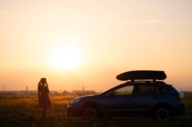 Yalnız kadının karanlık silueti çimenli çayırda arabasının yanında dinleniyor renkli gündoğumunun tadını çıkarıyor. Genç bayan sürücü SUV aracının yanında dinleniyor..