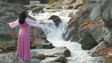 Uzun, modaya uygun bir gece elbisesi giymiş genç bir kadın ellerini kaldırıyor ve hızlı akan suyla küçük bir dağ nehrinin kenarında duruyor..