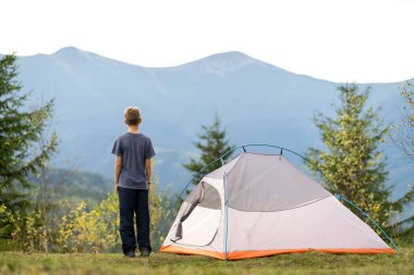 Kamp alanında çadırın yanında duran yürüyüşçü çocuk doğanın tadını çıkarıyor..