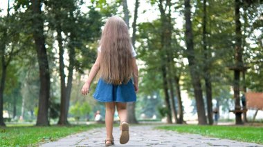 Yeşil parkta tek başına yürüyen yazlık elbiseli küçük bir kızın arka görüntüsü..