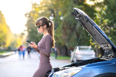 Kaputu açık, bozuk bir arabanın yanında cep telefonuyla konuşan genç üzgün bir kadın sürücü şehir caddesinde aracıyla sorun yaşaması için yardım bekliyor..