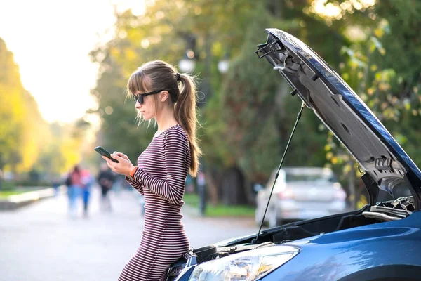 Kaputu açık, bozuk bir arabanın yanında cep telefonuyla konuşan genç üzgün bir kadın sürücü şehir caddesinde aracıyla sorun yaşaması için yardım bekliyor..