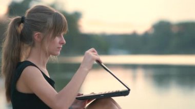 Laptopun arkasında çalışan genç bir kadın dışarıda, yaz parkında oturuyor..