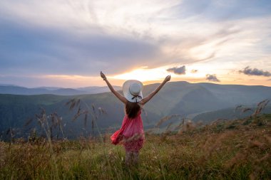 Kırmızı elbiseli mutlu bir kadın yürüyüşçünün arka görüntüsü çimenli bir tepede rüzgarlı bir sonbahar akşamında kollarını açıp güneşin batışının keyfini çıkarırken..