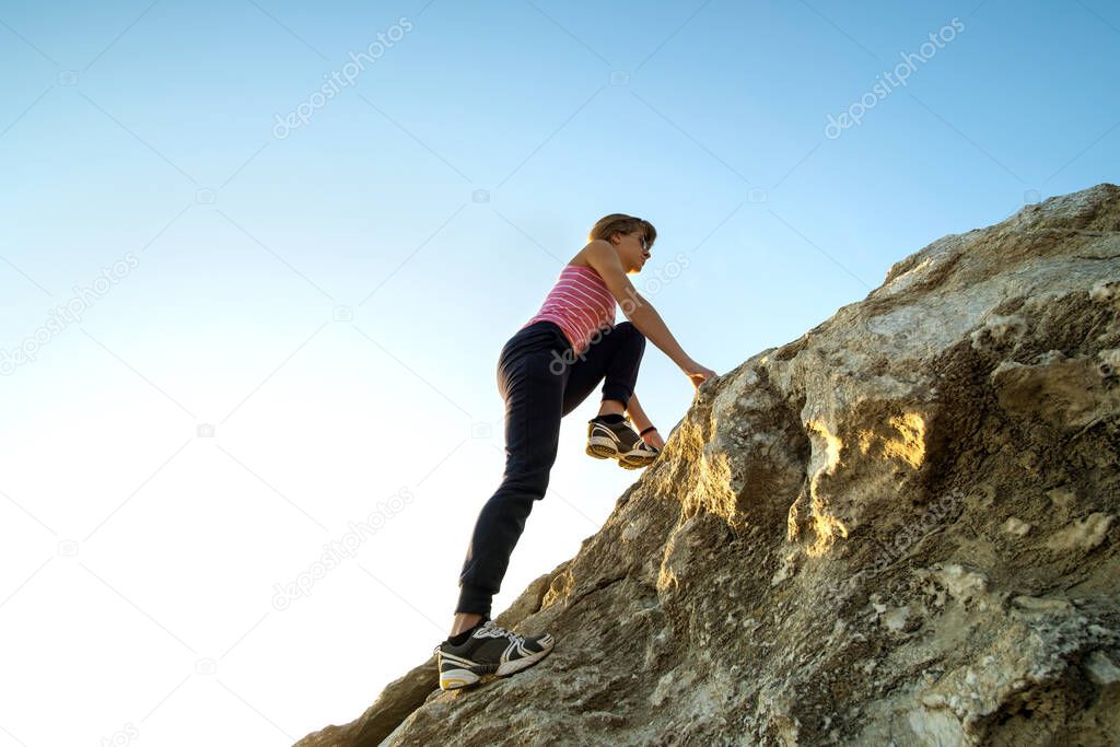 Woman hiker climbing steep big rock on a sunny day. Young female climber overcomes difficult climbing route. Active recreation in nature concept.