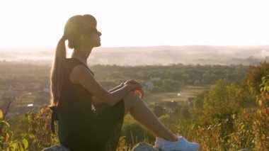 Siyah kısa elbiseli genç, sıska bir kadın yaz günbatımında bir kayanın üzerinde oturmuş dinleniyor. Modaya uygun bir kadın doğada sıcak bir akşamın tadını çıkarıyor..