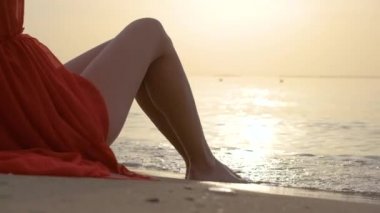 Kırmızı elbiseli, çıplak bacaklı genç bir kadın kumlu sahilde dinleniyor sıcak tropikal sabahın tadını çıkarıyor..