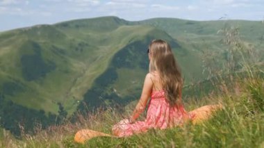 Kırmızılı genç kadın çimenli bir tarlada oturuyor rüzgarlı bir yaz gününde, dağlarda doğa manzarasının tadını çıkarıyor..