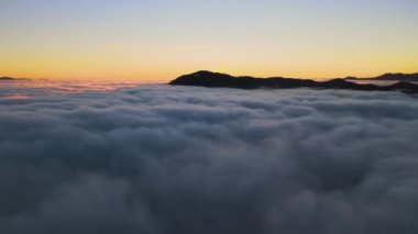 Ufuktaki uzak karanlık dağlarla beyaz yoğun bulutların üzerinde canlı gündoğumunun havadan görüntüsü..