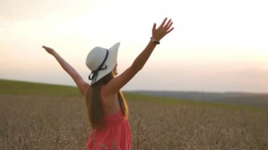 Kırmızı yaz elbisesi ve hasır şapkasıyla, olgun altın buğdaylı sarı çiftlik çayırında ellerini kaldırıp sıcak akşamın tadını çıkaran genç mutlu kadının arka görüntüsü..