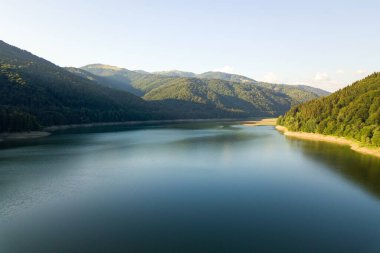 Yoğun yeşil ormanlarla kaplı yüksek dağ tepeleri arasında berrak mavi suyu olan büyük göl manzarası..