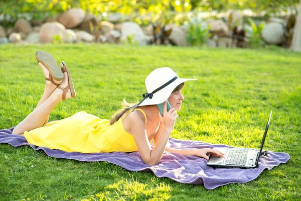 Girl student in yellow summer dress resting on green lawn in summer park studying on computer laptop having conversation on mobile cell phone. Doing business and learning during quarantine concept.