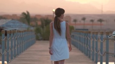 Beyaz elbiseli genç bir kadın uzun rıhtımda yürüyüşe çıkıyor. Akşamları dağ zirveleri siluetiyle. Tatil ve seyahat kavramı.