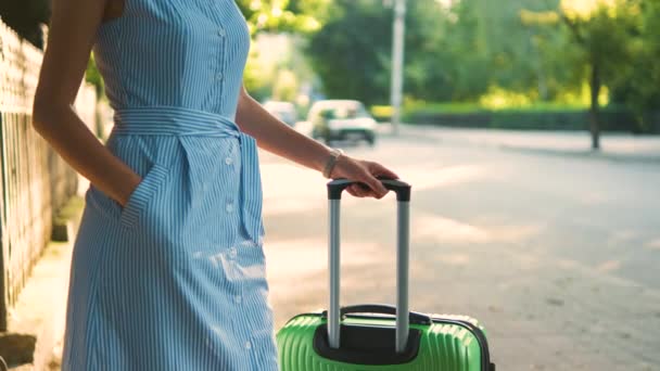 夏の日に緑のスーツケースを着た若い女性が街の歩道にタクシーが立っているのを待っています 旅行と休暇のコンセプト — ストック動画