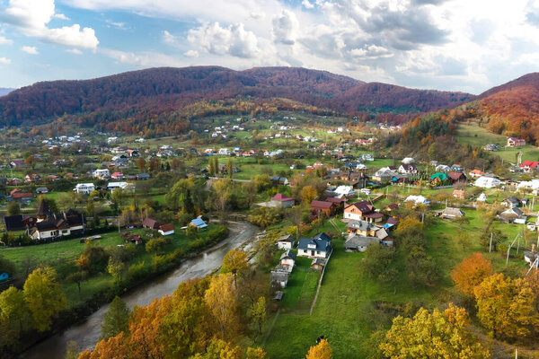 Aerial view of a village rural area with small houses between autumn mountain hills covered with yellow and green spruce forest.