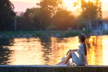 Gölün kıyısında tek başına oturan ve sıcak akşamın tadını çıkaran yalnız bir genç kadın. Doğada refah ve rahatlama kavramı.