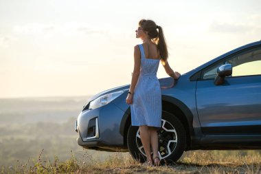 Mavi yazlık elbiseli mutlu kadın sürücü arabasının yanında sıcak bir akşamın tadını çıkarıyor. Seyahat ve tatil kavramı.