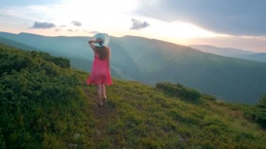 Kırmızılı genç kadın yaz dağlarında rüzgarlı bir akşamda yamaçtaki çayırda yürüyor..