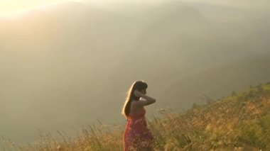 Kırmızılı genç kadın sonbahar dağlarında rüzgarlı bir akşamda çimlerde yürüyor..