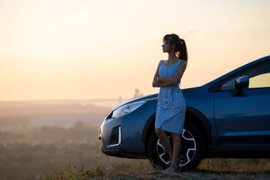 Mavi yazlık elbiseli mutlu kadın sürücü arabasının yanında sıcak bir akşamın tadını çıkarıyor. Seyahat ve tatil kavramı.