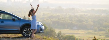 Genç bayan sürücü sıcak yaz akşamında arabasının yanında dinleniyor ve ellerini kaldırıyor. Seyahat hedefleri kavramı.