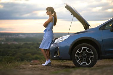 Kaputu açık, bozuk bir arabanın yanında duran genç bir kadın sürücü aracın motorunu kontrol ediyor ve yardım bekliyor..