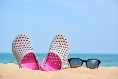 Sıcak, güneşli bir günde tropik deniz kıyısındaki kumlu kumsalda takunya ayakkabıları ve siyah koruyucu güneş gözlükleri. Yaz tatili kavramı.