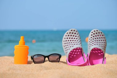 Sıcak güneşli bir günde, tropik deniz kıyısında takunya ayakkabıları, güneş kremi ve siyah koruyucu güneş gözlüklerine yakın çekim. Yaz tatili kavramı.