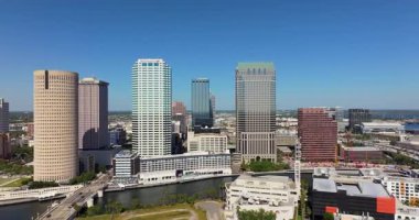 Tampa şehir merkezinin panoramik manzarası yükselmiş. Ticari yüksek binalar, köprüler ve güneşli gökyüzünün altında yeşil alanlar var.