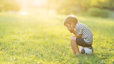 Küçük bir çocuk futbol topunun üzerine oturur ve çimenli bir arazide güneşli bir parıltıyla üzülür..