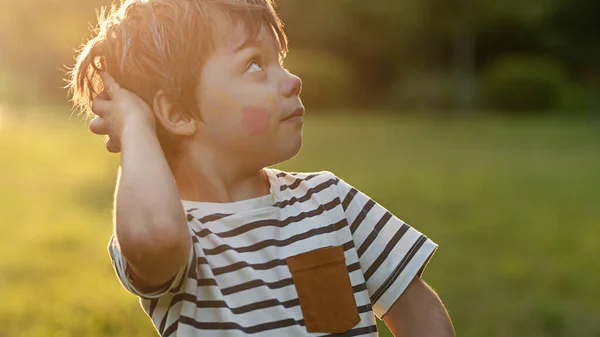 Niño con la cara pintada, mira sorpresa a un lado, en el verano en el parque —  Fotos de Stock