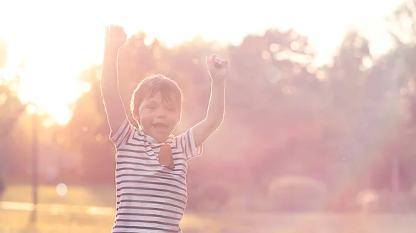 Chłopiec w świetle zachodu słońca w parku cieszy się życiem podnosząc ręce — Zdjęcie stockowe