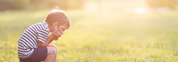 A little boy sits and is sad on a grassy field with a sun glare. — Stock Photo, Image