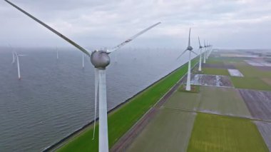 Bulutlu gökyüzünün altında deniz, hendek ve tarım arazileri etrafında dönen rüzgar türbinlerinin hava görüntüsü