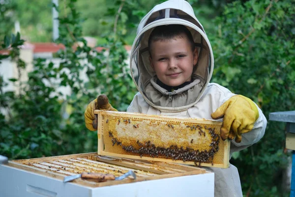 養蜂場の近くの保護服の中で陽気な少年養蜂家 蜂蜜とハニカム 有機食品の概念 最も役に立つ有機蜂蜜 — ストック写真