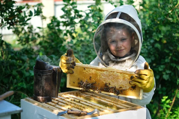 養蜂場の近くの保護服の中で陽気な少年養蜂家 蜂蜜とハニカム 有機食品の概念 最も有用な有機蜂蜜 子供たちを養蜂に惹きつける — ストック写真
