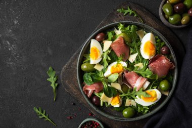Parmesanlı Prosciutto salatası, zeytin, yumurta ve roka siyah arka planda kesilmiş bir tabakta. İtalyan yemeği. Kopyalama alanı olan üst görünüm