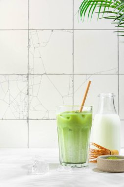 Beyaz fayans zemininde, sert gölgeler ve palmiye yapraklarıyla kaplı bir bardakta buzlu yeşil kibrit latte. Soğuk yaz içkisi. Dikey yönelim
