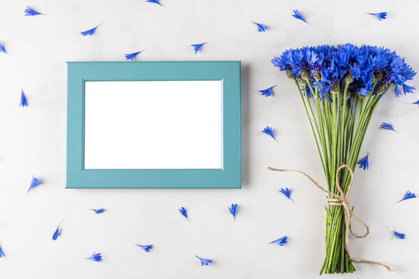 Blank photo frame with summer blue cornflowers bouquet on white marble background. top view. flat lay. mock up. festive holiday concept