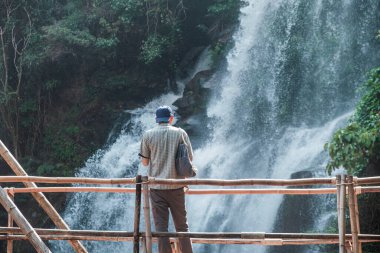 Rear view of man tourist standing on bamboo bridge facing majestic waterfall in lush tropical forest at Doi Inthanon National Park, Chiang Mai, Thailand. Eco-tourism and sustainable travel.