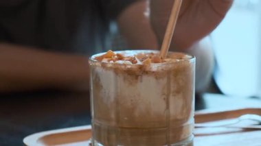Man mixing iced creamy coffee with straw on wooden tray in cozy cafe settings. Coffee culture, modern cafe lifestyle, artisan drinks and barista artistry, dessert beverage.