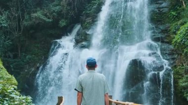 Man tourist standing on bamboo walkway facing majestic waterfall in lush tropical forest at Doi Inthanon National Park, Chiang Mai, Thailand. Vertical orientation. Eco-tourism and sustainable travel.