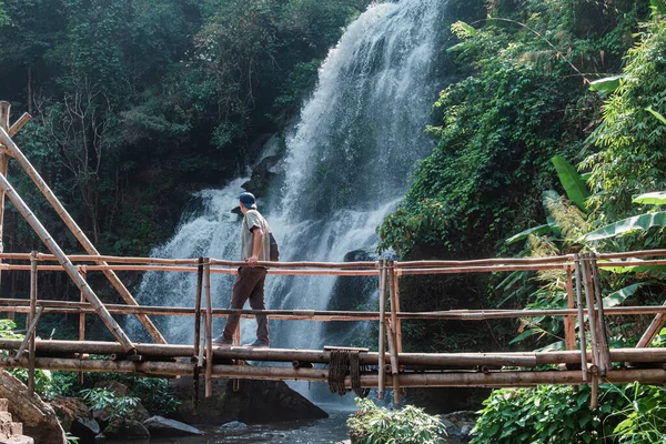 Man tourist walking on bamboo bridge across river with waterfall in lush tropical forest at Doi Inthanon National Park, Chiang Mai, Thailand. Eco-tourism and sustainable travel.