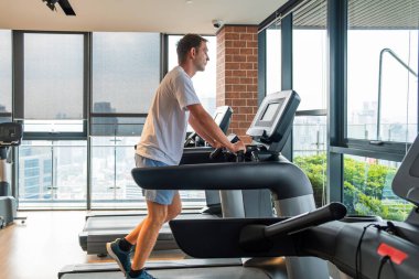 Şehir manzaralı modern bir spor salonunda koşu bandında egzersiz yapan bir adam. Erkek kardiyo yapıyor, spor eğitimi için ısınıyor ve konut binasındaki spor salonunda koşu bandında koşuyor. Zindelik, sağlık