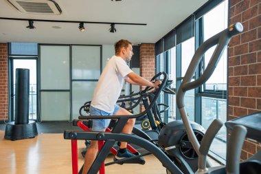 Modern spor salonunda şehir manzaralı eliptik bir antrenörde çalışan bir adam. Erkek, konut binasındaki fitness stüdyosunda sabit egzersiz bisikleti üzerinde kardiyo eğitimi alıyor. Zindelik, sağlık