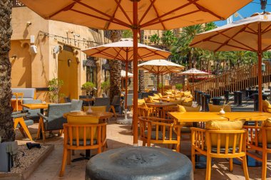 Madinat Jumeirah, Dubai, BAE 'de palmiye ağaçlarıyla çevrili ahşap sandalyeleri, masaları ve şemsiyeleri olan güneşli açık hava kafe terası. Yaz restoranı. Birleşik Arap Emirlikleri 'nde seyahat hedefi.