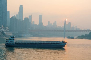Chongqing nehri, gün batımında kargo gemisi, kablo köprü olarak kaldı, Yangtze nehri üzerindeki yüksek binaların siluetleri, Çin. Nehir taşımacılığı ve şehir lojistiği. Nakliye, endüstri ve iç bölgeler
