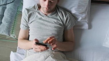 Adam akıllı telefon kullanarak yatakta yatıyor. Odaklanmış erkek uyandıktan sonra dinlenirken cihaza okur ve yazar. Modern dijital alışkanlıklar. Kişisel alandaki günlük teknoloji. Dijital çağda sabah rutini