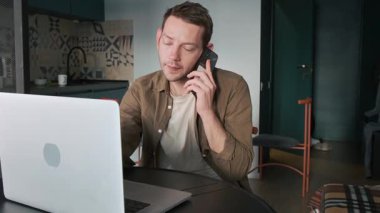 Ofis masasında dizüstü bilgisayarla çalışırken akıllı telefondan konuşan bir adam. Uzaktan çalışma, dijital bağlantı ve çevrimiçi ve telefon konuşmaları yoluyla görevleri yönetme konusunda erkek dijital göçebe. İş dünyası